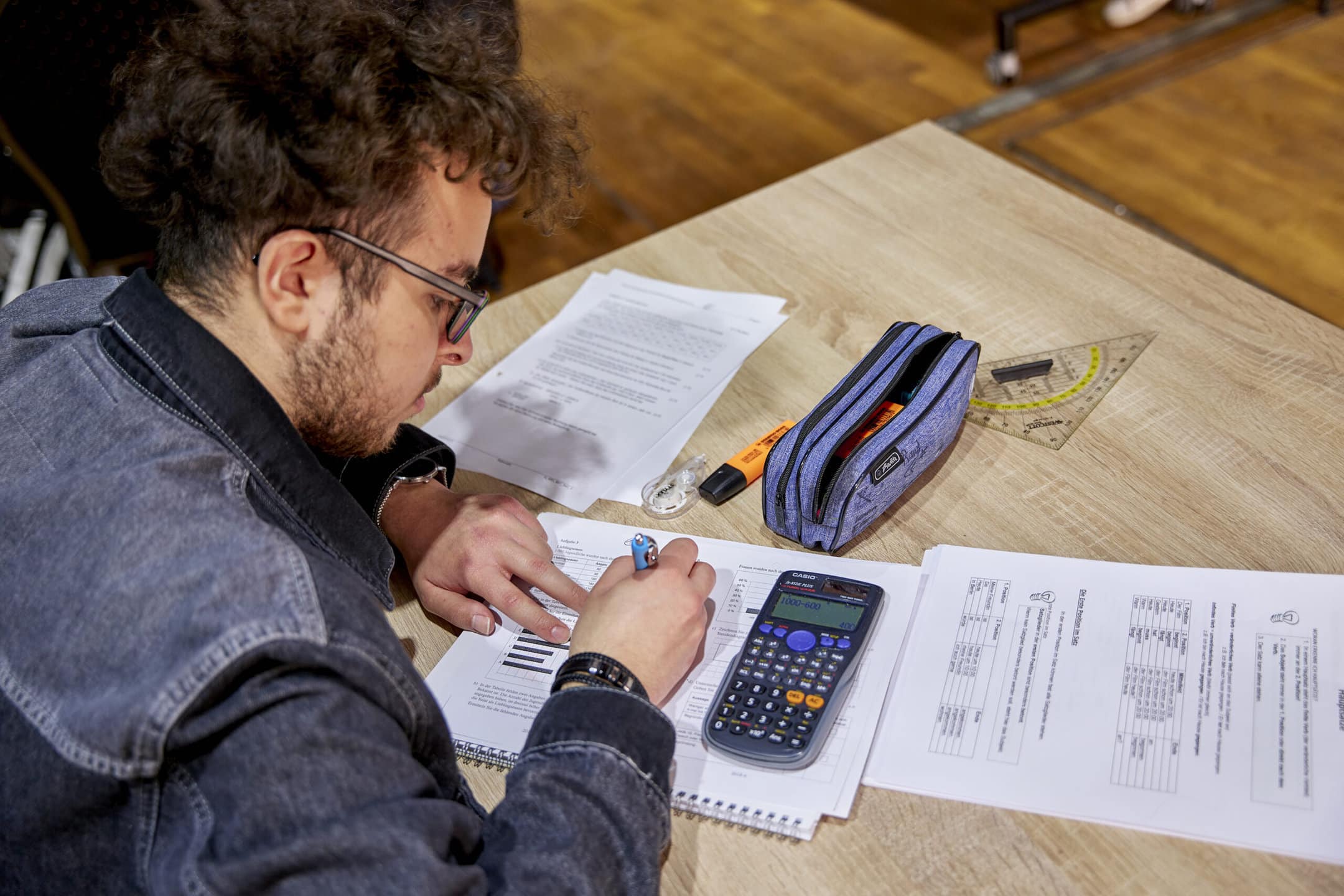 Ein Schüler sitzt an einem Tisch und arbeitet konzentriert an einer Rechenaufgabe, vor ihm liegen Arbeitsblätter, Schreibutensilien und ein Taschenrechner