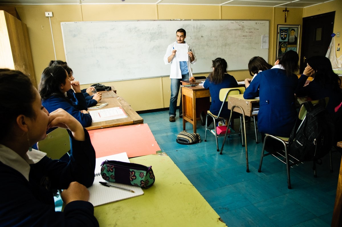 Fotografie aus einem Klassenzimmer, in dem mehrere Schüler:innen an Einzeltischen sitzen und ihren Blick nach vorne gerichtet haben, wo Gustavo Rojas Ayala ein Blatt Papier hoch hält