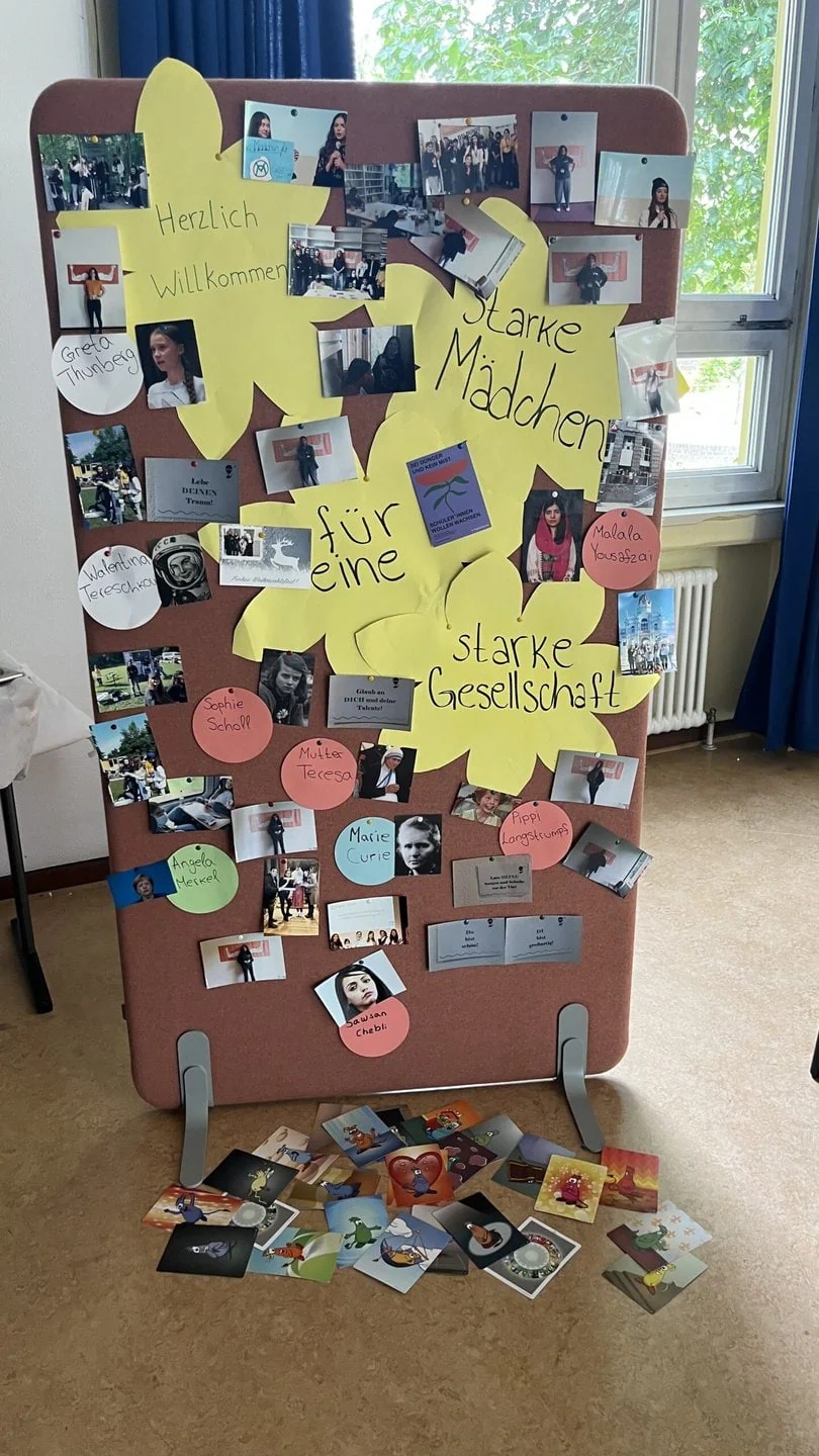Flipchart mit vielen Bildern und Fotografien daran gepinnt, dazwischen auf großen, blumenförmigen Schildern die Worte „Herzlich willkommen! Starke Mädchen für eine starke Gesellschaft“
