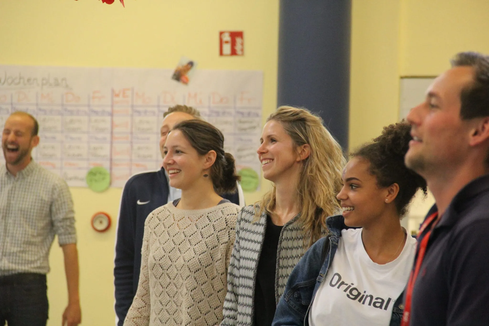 Bild von sechs lachenden Personen, die in einer Reihe in einem Workshopraum stehen; im Fokus ist eine blonde Frau