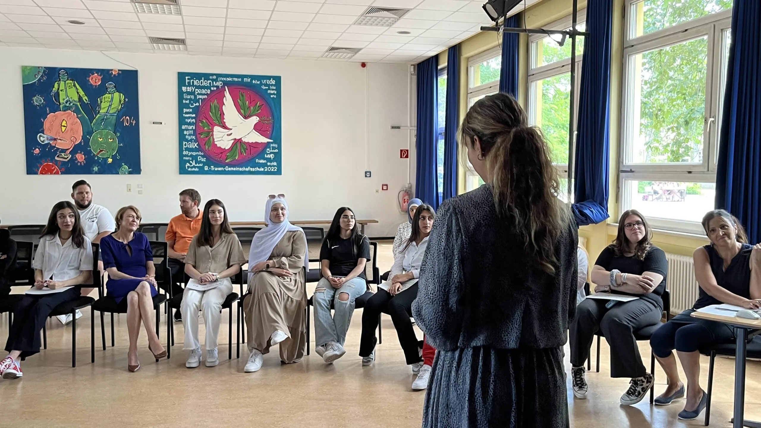 Eine Person steht in der Mitte des Stuhlkreises, im Hintergrund sitzen weitere Schüler:innen und Erwachsene – unter anderem Schirmherrin Elke Büdenbender – und hören zu.