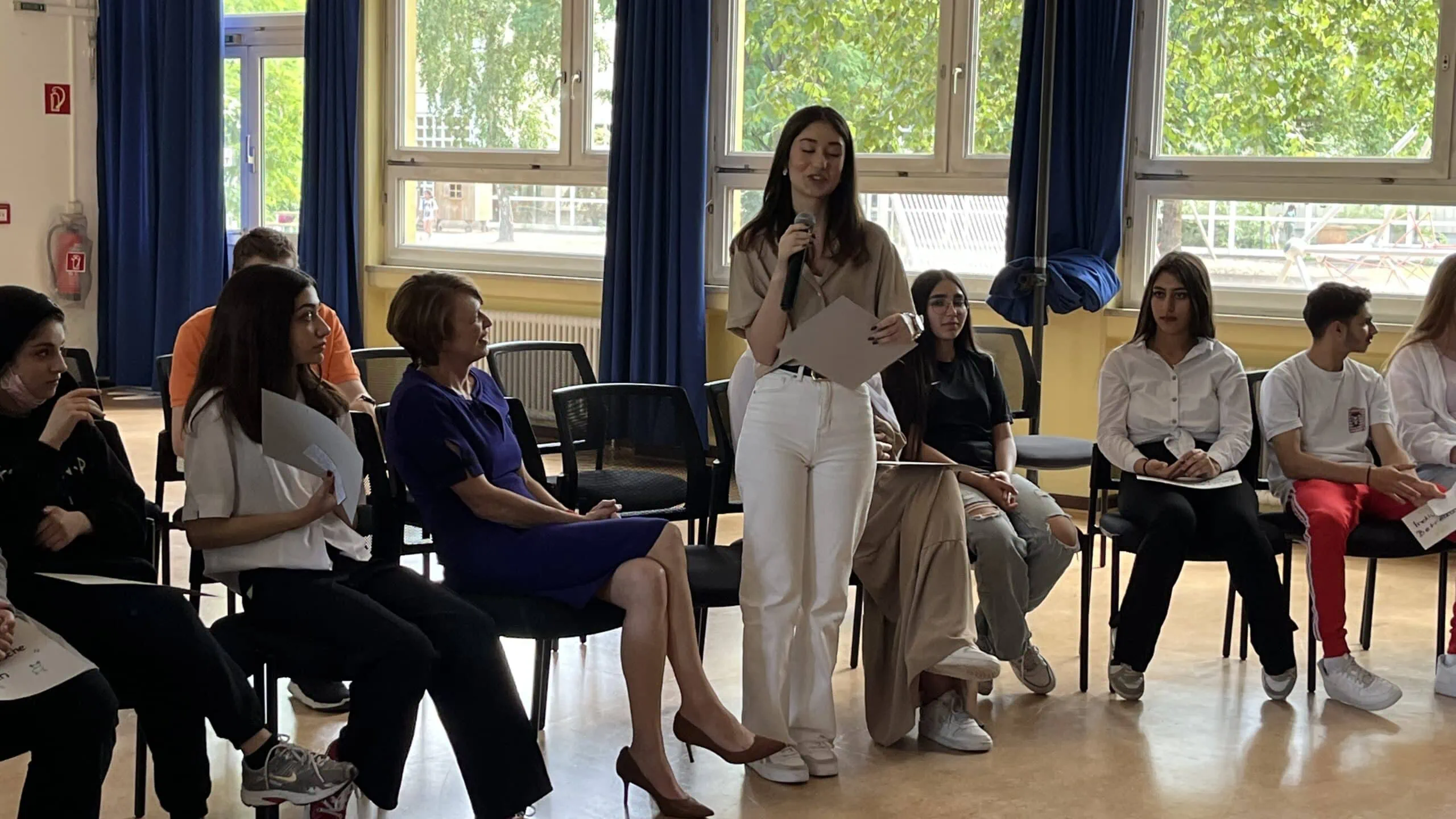 Eine Schülerin steht vor ihrem Stuhl im Stuhlkreis und spricht in ein Mikrofon, während die anderen Schüler:innen im Kreis sowie Schirmherrin Elke Büdenbender ihr zuhören.