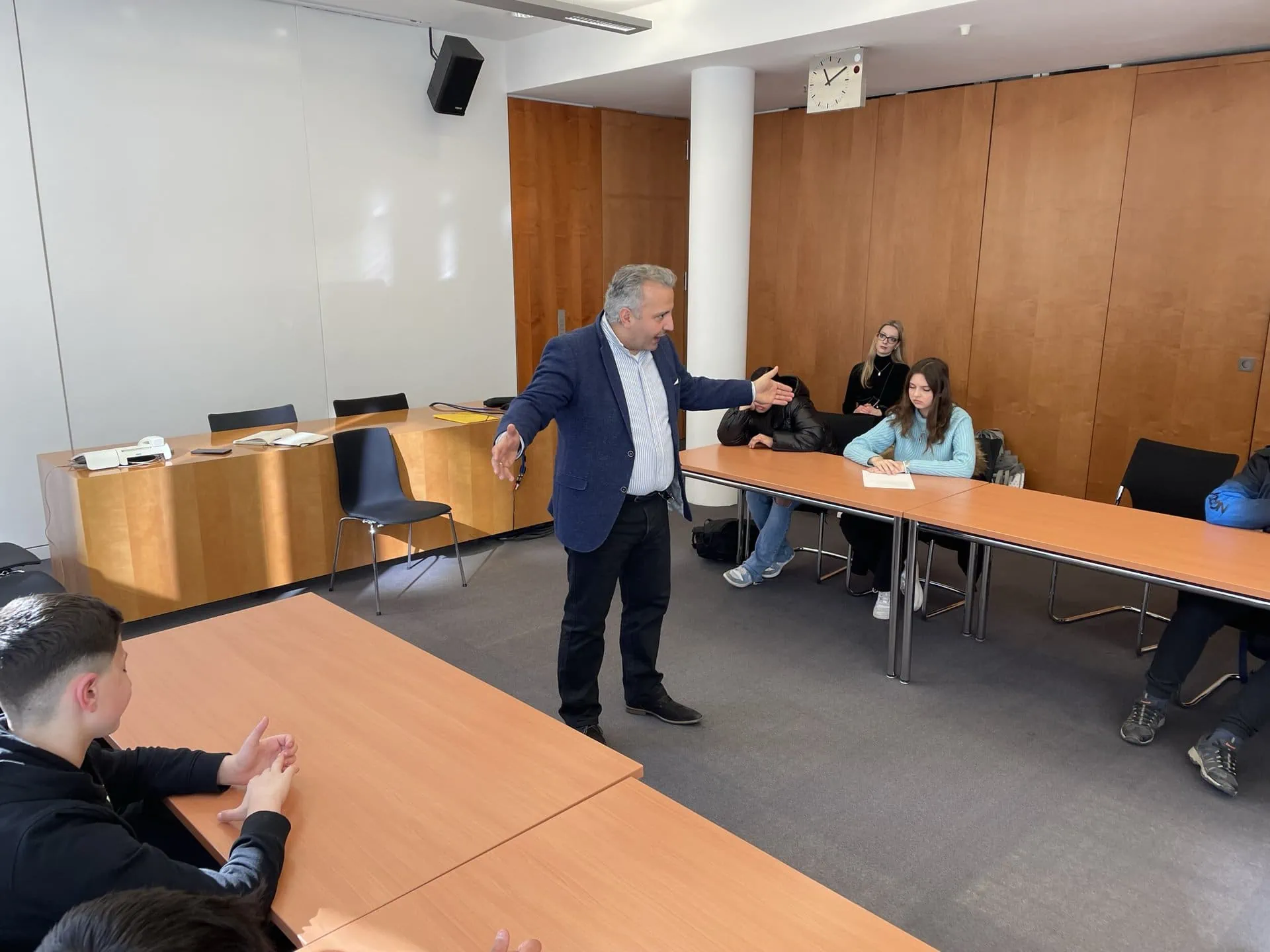 Abgeordneter des Reichstags in der Mitte eines Konferenzraumes, während er mit den Armen gestikulierend redet und an Tischen um ihn herum Schüler:innen an Tischen sitzen und zuhören