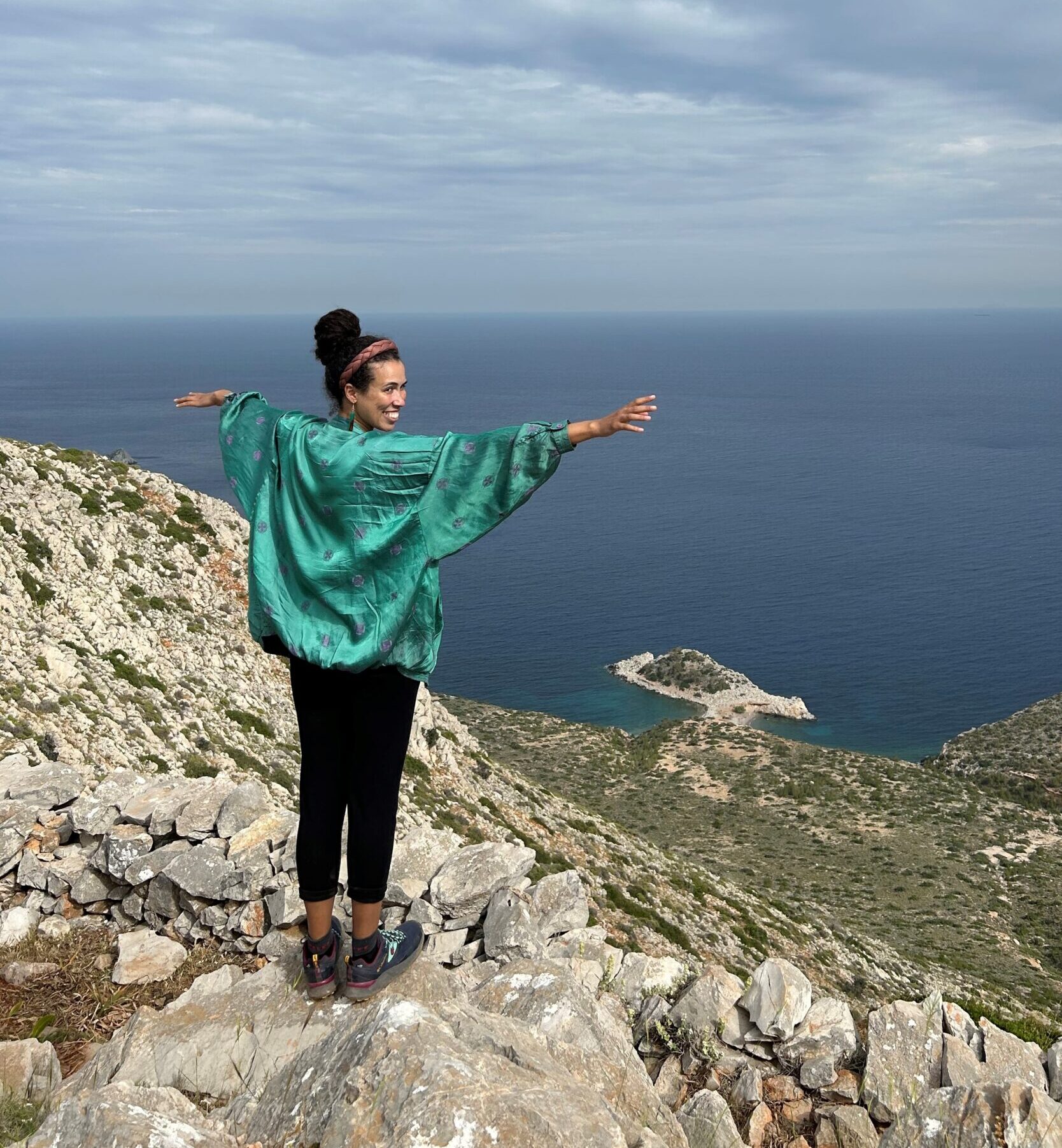 Foto von Natalie Rappert, die in einem grünen Oberteil auf einer klippe steht, die Arme weit ausgebreitet und den Kopf hinter sich in die Kamera dreht. Hinter ihr ist eine felsige Küste, das Meer und wolkiger Himmel zu sehen.