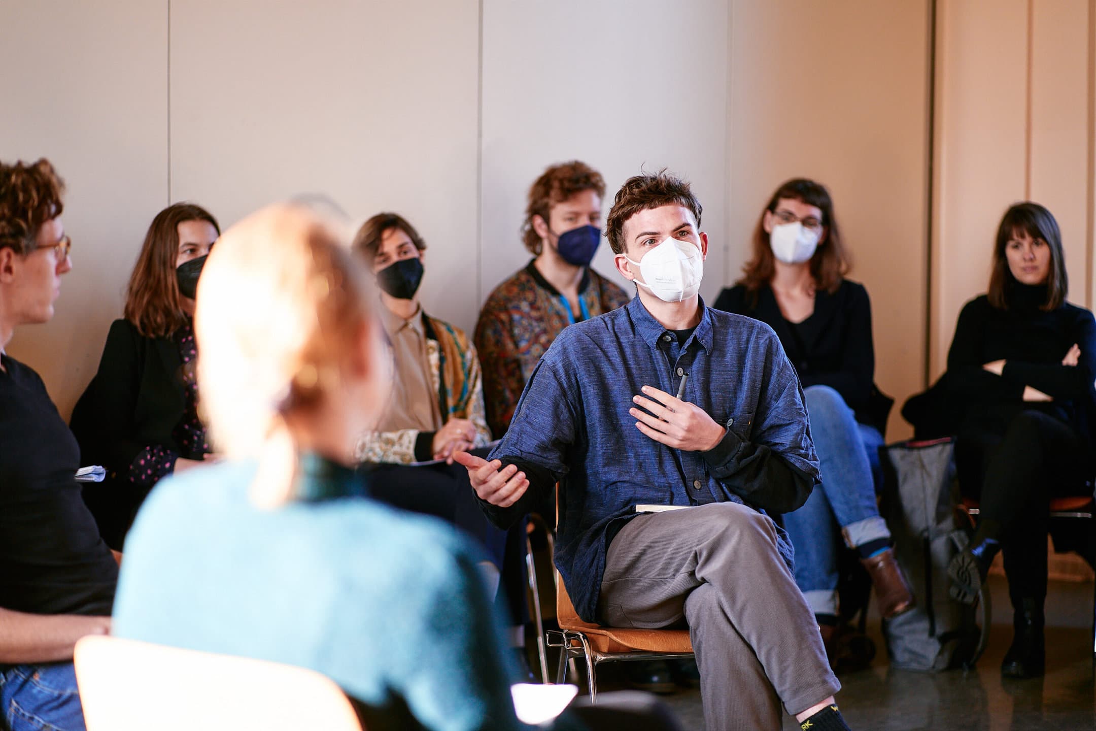 Fotografie der Gesprächsrunde zum Quereinstieg in der Schule am Schillerpark Berlin. Im Fokus ist ein Berliner Fellow, der eine weie FFP2-Maske trägt, und redend gestikuliert. Im Vor- und Hintergrund sitzen weitere Personen, die Masken tragen und aufmerksam zuhören.