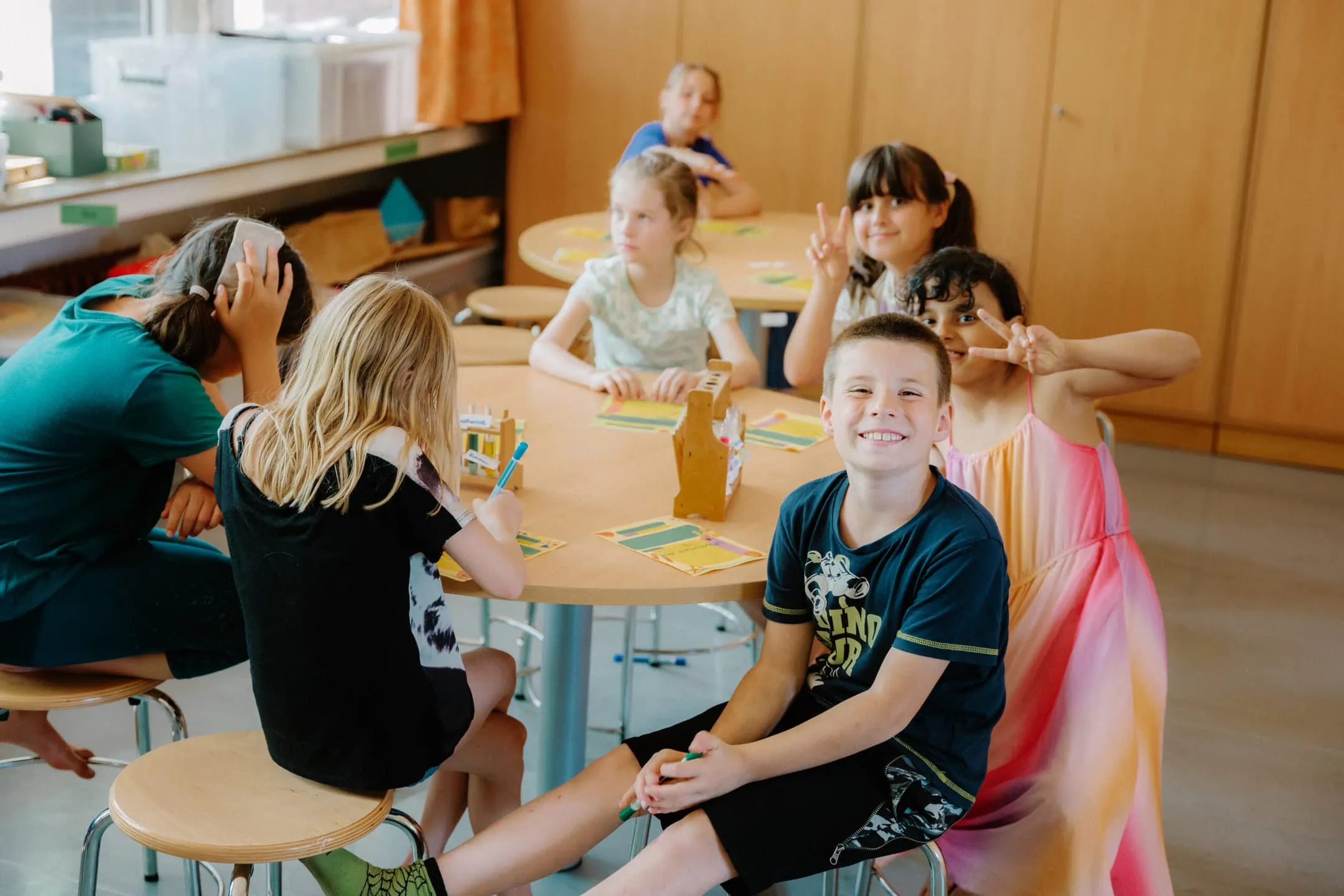 Fünf Schüler:innen sitzen in einem Klassenzimmer an einem runden Tisch, drei von ihnen schauen in die Kamera. Im Fokus ist ein Junge, der breit ins Bild grinst, die zwei Mädchen hinter ihm zeigen das Peace-Zeichen hoch.