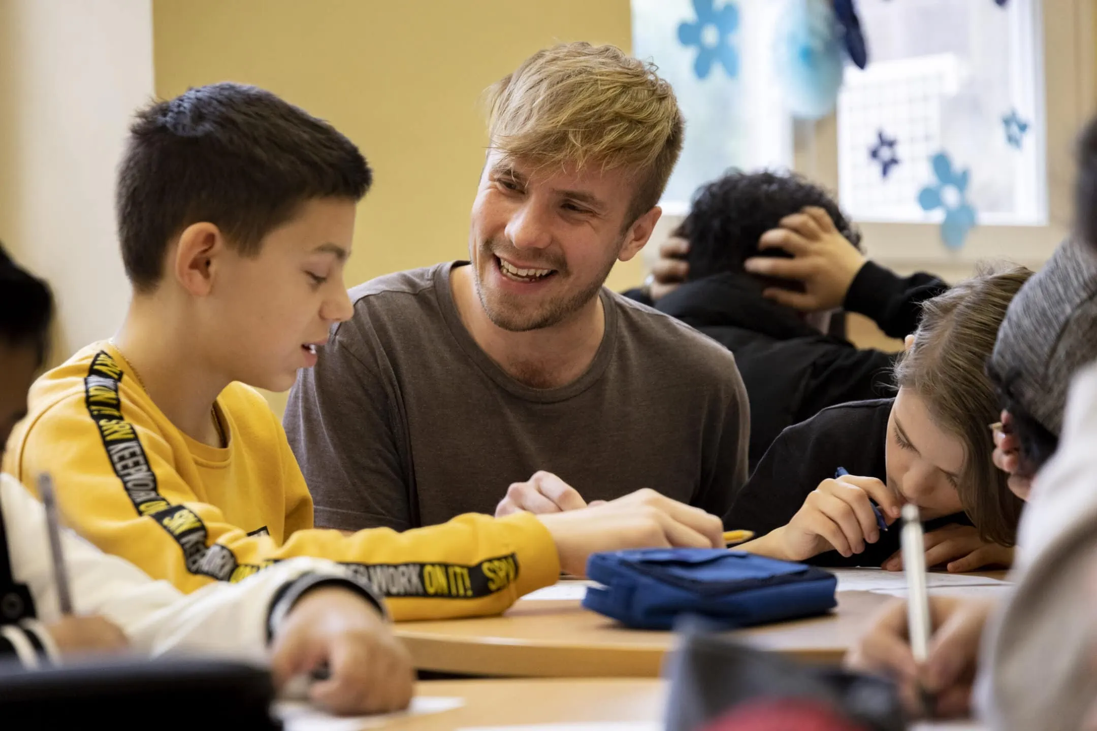 Süd Fellow 2022 Janosch Warda im Unterricht mit einem Schüler. Janosch kniet neben dem Schüler, der am Tisch sitzt, und lacht ihn an.