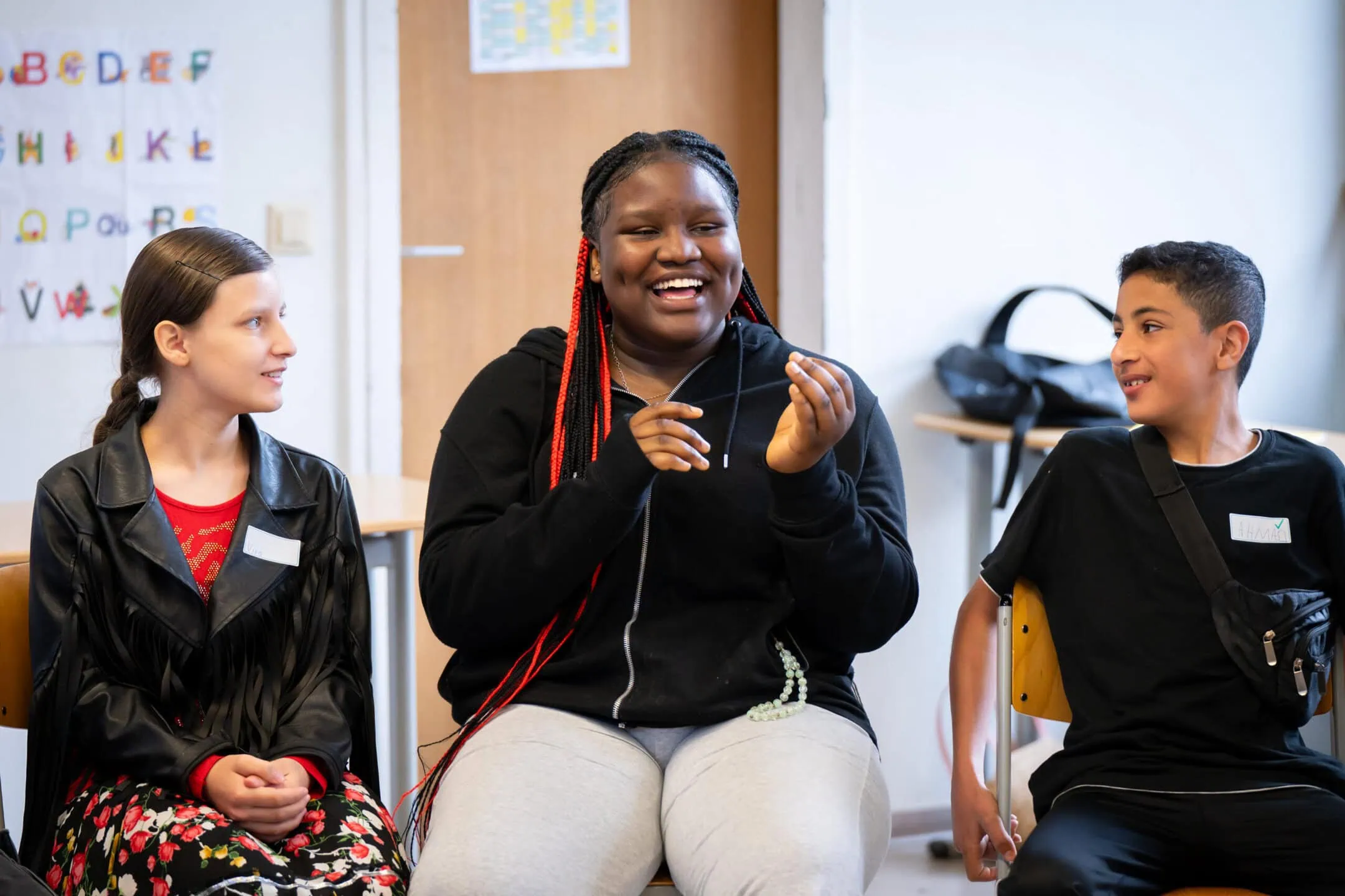 Drei Kinder sitzen in einem Klassenzimmer, lächeln und reden miteinander.